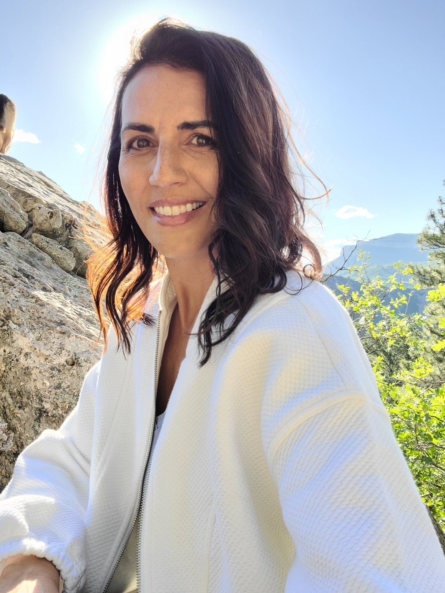 Toni smiling on a sunlit mountain trail in a cream cardigan